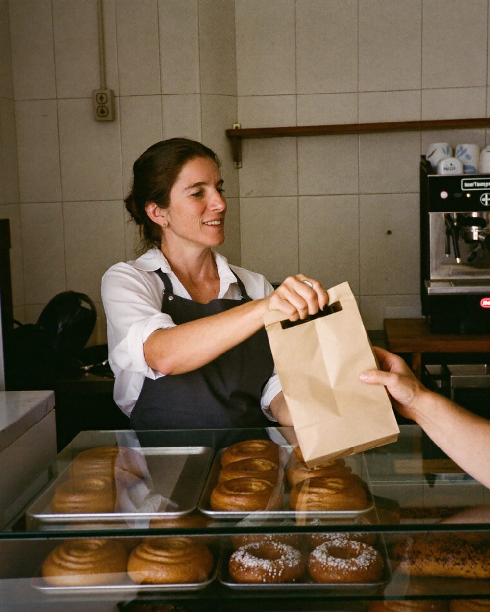 Dona de uma pastelaria a entregar um saco de pão a um cliente, ao balcão